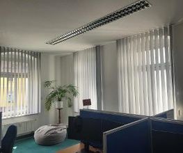 Schulungsraum mit 3 Fenstern und Vorhängen. Eine Pflanze steht vor einem Sitzsack. Rechts sind mehrere Arbeitsplätze erkennbar.