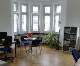 Heller Raum mit einigen Arbeitsplätzen sowie einer Sitzgelegenheit bestehend aus einem runden Tisch, auf dem Blumen stehen, und 4 Stühlen.