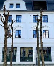 Außenansicht (Frontalansicht) des weißen, klassisch-modernen Gebäudes mit schwarz gerahmten Fenstern. Im Vordergrund Bäume.