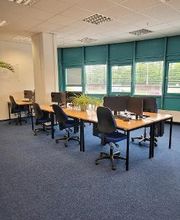 Ein Schulungsraum mit mehreren Arbeitsbereichen. Jeder Arbeitsplatz besteht aus 2 PC-Bildschirmen, Tastatur und Maus sowie Headset und einem blauen Bürostuhl. Türkise Fensterfront