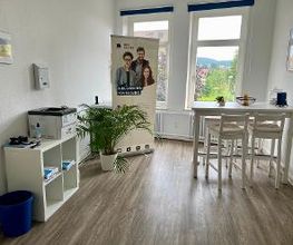 ein heller Aufenthaltsraum mir 2 großen Fenstern. Rechts steht ein weißer Tisch mit Bar-Hockern. Links steht ein Regal, auf dem ein Drucker steht. Daneben steht eine Zimmerpflanze und ein Roll Up der WBS GRUPPE