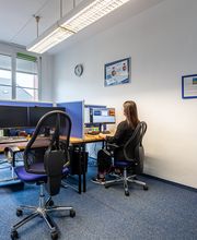 Raum mit mehreren Arbeitsplätzen mit jeweils 2 Bildschirmen, einem Bürostuhl, Tastatur und Maus. Am rechten Platz sitzt eine Frau und arbeitet. An der Wand hängen Plakate und eine Uhr. Der Boden ist mit blauem Teppich ausgelegt.