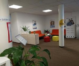 Gemütliche Lobby mit Sitzbereich bestehend aus 2 roten Sofas, und Sitzhocker. Werbeaufsteller, Plakate an der Wand und einige, grüne Zimmerpflanzen.