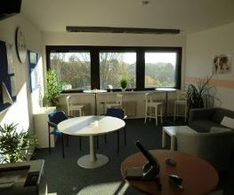 Pausenbereich mit3 Fenstern und Blick ins Grüne. Davor 2 Tische mit jeweils 2 Barhockern, einem größeren Tisch mit 2 Stühlen und einem grauen Sofa. An der Wand mehrere Pinnwände und Bilder. Ein Drucker steht im Raum