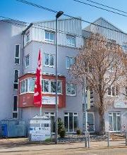 Dieses Bild zeigt das Gebäude auf der Carl-Benz-Straße von außen. Das Haus hat eine hellgraue Fassade und vor dem Gebäude stehen 2 Bäume sowie eine rote Fahne.