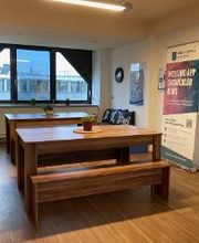 Pausenraum mit 32 Tischen und 4 Bänken. Hinten im Raum ein Sofa, daneben ein Roll Up Banner der WBS CODING SCHOOL.