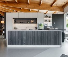 Diese moderne LEICHT Designküche verbindet klare Linien, hochwertige Materialien und intelligente Funktionalität zu einem stilvollen Gesamtkonzept. Die großzügige Kücheninsel mit grauer Holzstruktur bildet den Mittelpunkt der offenen Wohnküche und bietet viel Arbeits- und Stauraum. Helle Hochschränke, integrierte Elektrogeräte sowie offene Regale mit indirekter LED-Beleuchtung schaffen eine elegante und wohnliche Atmosphäre. Die Ausstellungsküche im Küchentreff Liebold zeigt zeitloses Küchendesign für höchste Ansprüche an Ästhetik und Komfort.