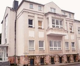 gelbes Gebäude mit mehreren Fenstern und einem gläsernen Eingang auf der linken Seite mit Treppe.