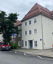 Außenansicht des Standorts Weilheim. Dreistöckiges Gebäude mit Spitzdach. Davor eine Einbahnstraße mit geparkten Autos.