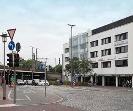 Eine Straßenecke mit einem weißen mehrstöckigen Gebäude, das Büros und Geschäfte beherbergt, darunter „ver.di“. Links ist eine Unterführung zu sehen, vor der Autos warten. Es gibt Ampeln, Verkehrsschilder und einen Fahrradweg. Der Himmel ist bewölkt.