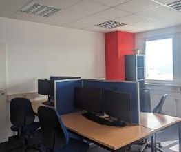 Schulungsraum mit mehreren Arbeitsbereichen, die durch blaue Trennwände voneinander abgegrenzt sind und mit PC-Bildschirmen, Tastatur, Maus und Headset ausgestattet sind. Blaue Bürostühle.