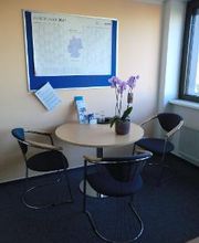 Bild von einer Sitzecke. 1 runder Tisch und 3 Stühle. Auf dem Tisch steht eine lavenderfarbene Topfpflanze und an der Wand hängt eine blaue Pinnwand an der ein Kalender befestigt ist. 