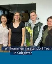 Team aus 4 Personen die freundlich in die Kamera schauen. Ein Kollege hält eine gelbe Orchidee, am unteren Bildrand der Text "Willkommen im Standort Team in Salzgitter"
