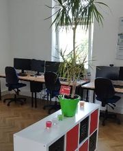 Schulungsraum mit mehreren Arbeitsbereichen, mit 2 PC-Bildschirmen, Tastatur, Maus und Headset ausgestattet sind. Im Vordergrund steht ein weißes Regal mit schwarzen Fächern und darauf steht eine große Zimmerpflanze sowie bunte Kerzen in Gläsern.