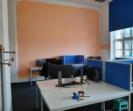 Arbeitsbereich mit 2 Computer-Bildschirmen, einer Tastatur, Maus und blauen Trennwänden. Auf dem Tisch liegen 2 Locher und links befindet sich ein Garderobenständer.