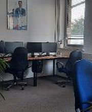 Schulungsraum mit 4 Arbeitsplätzen mit jeweils 2 Bildschirmen, einem Bürostuhl, Tastatur und Maus. Mehrere Pflanzen stehen im Raum, sowie ein Ventilator.