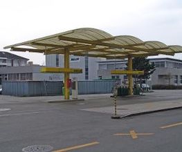 AGROLA Tankstelle in St. Margrethen, Sankt Margrethen; geschwungene Überdachung mit gelbem Gerüst. Im Hintergrund Gebäude mit grauer Fassade.