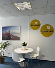 Gemütliche Sitzecke mit einem runden, weißen Tisch und 3 Stühlen. An der Wand hängt ein Bild von einem Cabrio mit Surfbrett am Strand und 2 gelbe, runde Aufkleber auf denen steht Herzlich Willkommen und Wir gestalten gemeinsam.