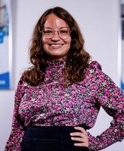 Bild einer Mitarbeiterin. brünett, gewellte Haare, Brille, Bluse mit Blumenmuster, freundliches Lächeln.