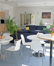 Helle, voll ausgestattete Teilnehmerküche mit Getränkeautomaten. 2 blaue Sofas und weiße Tische mit Stühlen. Mehrere Zimmerpflanzen und ein Bild an der Wand.