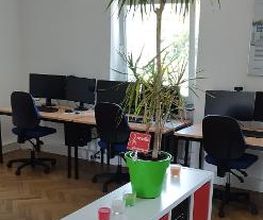 Schulungsraum mit mehreren Arbeitsbereichen, mit 2 PC-Bildschirmen, Tastatur, Maus und Headset ausgestattet sind. Im Vordergrund steht ein weißes Regal mit schwarzen Fächern und darauf steht eine große Zimmerpflanze sowie bunte Kerzen in Gläsern.