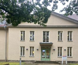 Außenansicht vom Gebäude. Helle Fassade mit weißen Fensterrahmen und einer grünen Eingangstür.