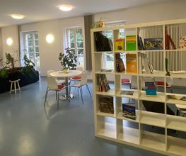 Aufenthaltsraum mit blauen Sofas, weißen Tischen und Stühlen, einem Bücherregal, großen Fenstern und einigen Zimmerpflanzen.
