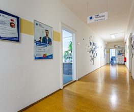 Flur mit mehreren Plakaten an den Wänden und Türen die in verschiedene Zimmer führen. Am Ende des Flures steht ein Roll up Banner und an der Decke hängt ein Schild mit Wegpfeil zum Empfang.
