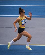 Eine Leichtathletin bewegt sich dynamisch über die Laufbahn in einer Wettkampfsituation. Die Körperhaltung zeigt Technik und hohe Konzentration.