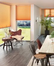 Ein moderner Essbereich mit bodentiefen Fenstern, die mit orangefarbenen Plissees ausgestattet sind. Die Plissees schaffen eine angenehme Lichtstimmung und unterstreichen die Raumgestaltung.