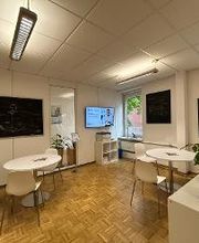 Ein gemütlicher Aufenthaltsraum mit weißen, runden Tischen an denen jeweils 2 weiße Stühle stehen. Es befinden sich einige Regale und Zimmerpflanzen in dem Raum und an den Wänden hängen kunstvolle Bilder von einem Anker und Leuchtturm