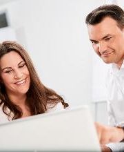 Dame, weiß gekleidet, dunkle lange Haare, sitzt am Tisch mit einem Notebook und lächelt ins Bild. Neben ihr sitzt ein Herr, dunkle Haare, weißes Hemd, und zeigt etwas am Notebook.