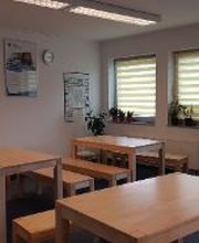 Gemütlicher Pausenraum mit hellbrauen Tischen und Bänken. An der Wand hängt ein gelbes Bild auf dem eine Kaffeetasse gemalt ist und das Wort Pause geschrieben steht. 