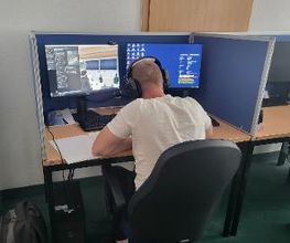 Mann sitzt am Arbeitsplatz mit 2 Bildschirmen und einer Tastatur. Er trägt ein Headset und vor ihm liegen mehrere Blätter. Rechts ein weiterer Arbeitsplatz.