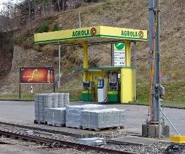 AGROLA Tankstelle in Mogelsberg; Im Vordergrund Zuggleise und Paletten mit Setzsteinen. AGROLA Tankstelle mit zwei Säulen und Zahlterminal. Totem mit Preisen im Hintergrund