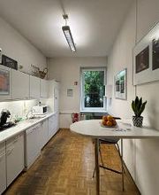 Eine Küche mit Kaffeemaschine, Wasserkocher und Mikrowelle. Auf dem Tisch befindet sich ein Teller mit Orangen sowie eine grüne Topfpflanze. Es hängen Bilder an der Wand.