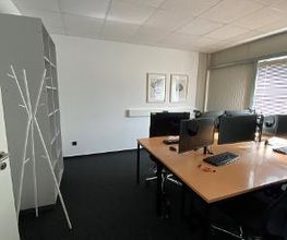 Schreibtische im Trainingsraum mit jeweils 2 PC-Bildschirmen, Tastatur, Headset, Maus und zwei Bildschirmen, Garderobe steht unmittelbar neben einem Bücherregal.