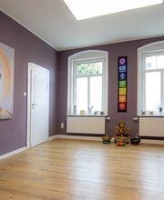 Ein geräumiger und ruhiger Meditations- und Entspannungsraum mit violetten Wänden und hellem Holzboden. Links an der Wand hängt ein großformatiges Gemälde eines Buddha-Kopfes in warmen Gold- und Beigetönen mit einem leuchtenden Heiligenschein. In der Mitte der hinteren Wand hängt eine bunte Chakra-Wanddekoration mit sieben farbigen Symbolen, die für die Energiezentren des Körpers stehen. Unterhalb der Dekoration stehen mehrere Buddha-Statuen, Klangschalen und Kerzen auf dem Boden. Die beiden Fenster lassen viel Tageslicht herein, und die schlichte Raumgestaltung fördert eine harmonische und entspannende Atmosphäre.