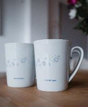 2 weiße Tassen mit Raketen drauf auf einem Tisch. Dahinter ein Strauß Blumen.