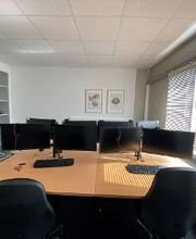Schreibtische im Trainingsraum mit zwei Bildschirmen, Tastatur, Maus und Headset. Neben den Schreibtischen steht ein Bücherregal und eine Graderobe. An der Wand hängen zwei Bilder