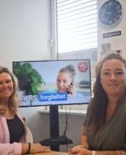 Beratungsteam mit viel Herz. 2 Frauen sitzen in einem Büro vor einem Bildschirm und blicken freundlich in die Kamera.