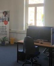 Schulungsraum mit mehreren Arbeitsplätzen jeweils mit 2 Bildschirmen, einem Bürostuhl, Tastatur, Maus und Headset. Im Hintergrund ein WBS TRAINING Roll Up Banner und eine Pflanze.