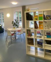 Aufenthaltsraum mit blauen Sofas, weißen Tischen und Stühlen, einem Bücherregal, großen Fenstern und einigen Zimmerpflanzen.