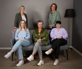 Eine Gruppe von 5 Frauen, die teilweise stehen und teilweise auf einer Couch sitzen.