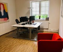 Hell, freundlich und modern gestalteter Beratungsraum mit zwei weißen Schreibtischen vor einem Fenster, auf dem Bildschirme und Tastaturen stehen. An den weißen Wänden Bilder. Im Vordergrund ein roter Lounge-Sessel. Parkettfußboden.