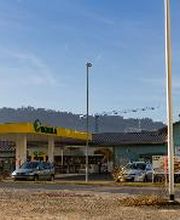 AGROLA Tankstelle in Reinach; links Tankstelle mit TopShop; grosse Überdachung; blaues Gebäude angeschlossen; rechts Mast im Vordergrund daneben Totem mit Landi Schild, AGROLA Schild und Preisanzeigen