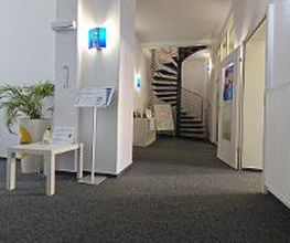 Empfangsbereich mit links einem Beistelltisch und Plakaten. Im Hintergrund ein Regal neben einer Wendeltreppe.