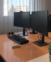 Arbeitsbereich mit 2 Computer-Bildschirmen, einer Tastatur, Maus und Headset. Fenster im Hintergrund und ein kleine Topfpflanze auf dem Tisch.