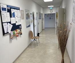Flurbereich mit Pinnwand und Plakaten an der Wand. Mehrere Türen zu Räumlichkeiten. Ein dekorativer Strauch aus Zweigen und ein Stuhl.