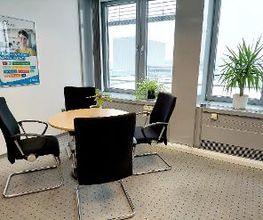 Raum mit einem Tisch und 4 schwarzen Stühlen. Links hängt ein Plakat an der Wand, rechts steht eine Zimmerpflanze auf dem Fensterbrett. Der Boden ist mit Teppich ausgelegt.